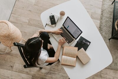 Overhead shot of a woman working on a computer with a book open next to her.