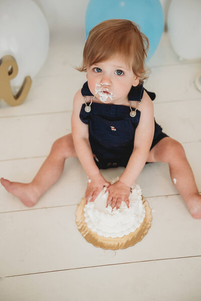 Baby boy looks up with cake on his face while eating birthday cake.