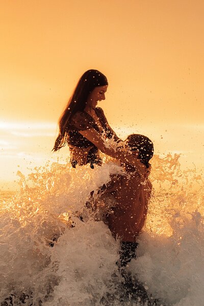 woman lifts on to man's shoulders as wave crashes on them and they laugh at sunset in wailea maui