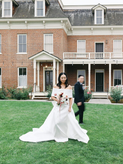 A bride in a flowing white gown holds flowers in front of the Barton House. A blurred groom in a suit walks by.