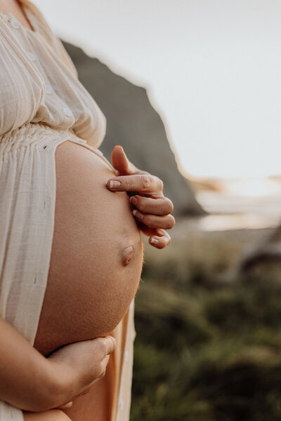 A mother touches her pregnant belly wearing a white dress at sunset in christchurch