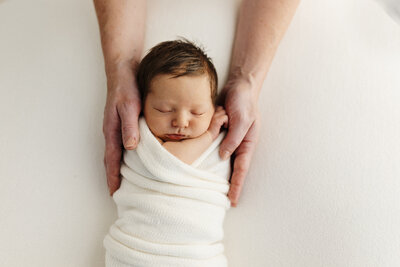 A little baby boy held in his parents hands, the image is in colour and the baby has lush brown hair