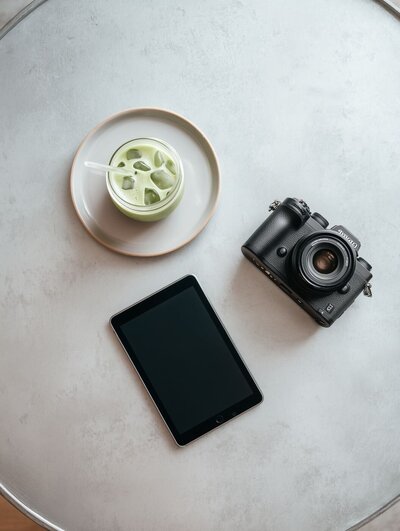 Flat lay of a creative workspace with an iced matcha latte, black digital camera and tablet on a light grey round table