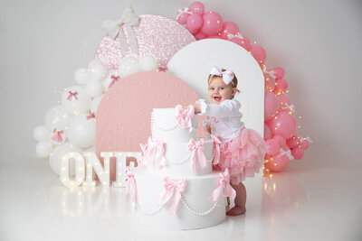 Toddler holding on to a giant cake prop with bows and arches in the background in pink and white.