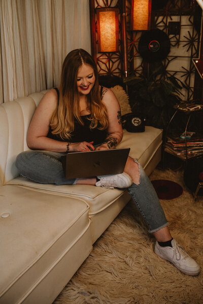 A blonde woman in jeans and a black top sits on a white couch and is smiling at her laptop.