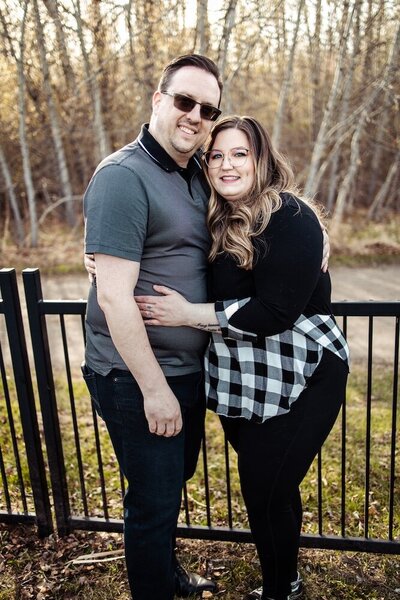 A man and a woman hug each other while standing in front of a black fence near the woods