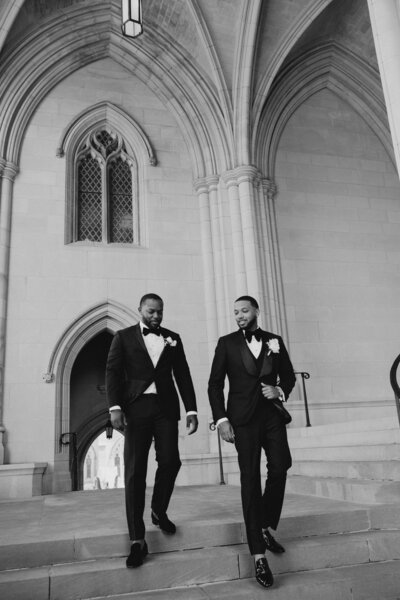 Two men in tuxedos walk down church steps in black & white