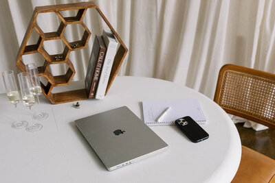 A round white table with a closed laptop, a smartphone, a notepad with a pen, and two champagne glasses. A hexagonal wooden shelf on the table holds two books. A woven chair is partially visible behind the table.