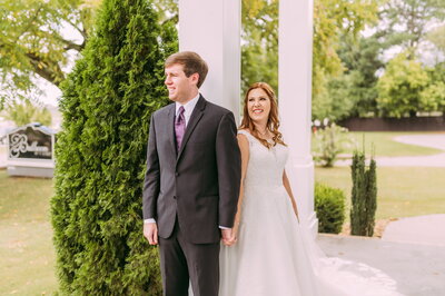 An image showing a Bride and Groom holding hands in front a column and a tree. They are smiling and looking off camera.