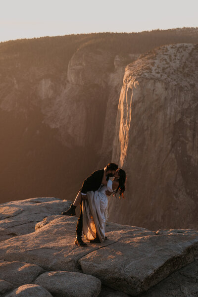 groom dips bride and holds her leg while they kiss on a rock cliff overlooking Yosemite valley with El Capitan in the background and the sun is shining on the rock behind them