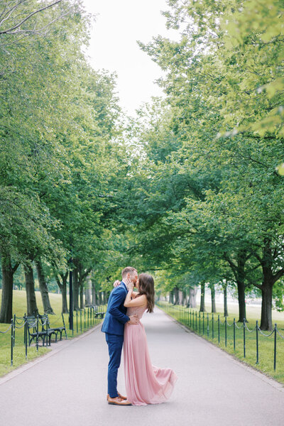 Engagement photo of a couple in downtown Washington, D.C., surrounded by lush greenery; the woman wears a flowing pink dress, the man a navy suit, as she wraps her hand around his head while they lean in for a kiss