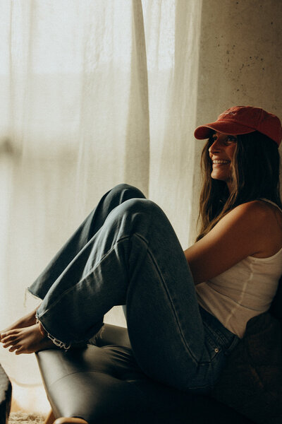 a woman in a red cap smiling while sitting in front of a sheer curtain for brand photography photoshoot on the Sunshine Coast