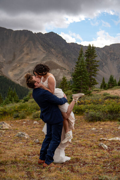 Groom lifts up his wife and kisses her as she kicks her feet up with the Colorado mountains in the background.