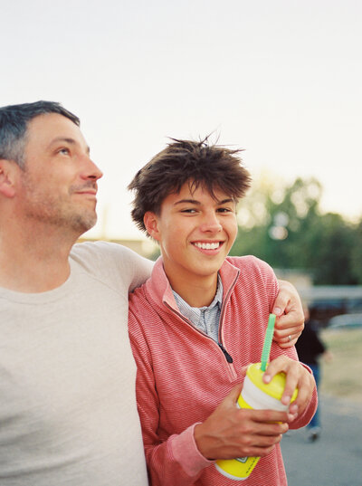 Teenage son with a lemonade in hand laughs while embraced by his father at a night out at the fair.