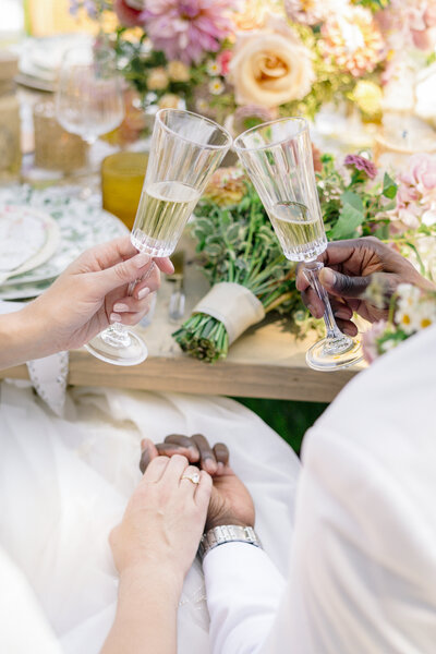 A couple clinks their champagne glasses in celebration, with a bouquet of flowers and a decorative table setting in the background. Their hands are gently clasped together on the table.