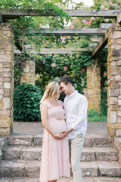 A couple stands under an arbor in a garden during their New Jersey Maternity photos.