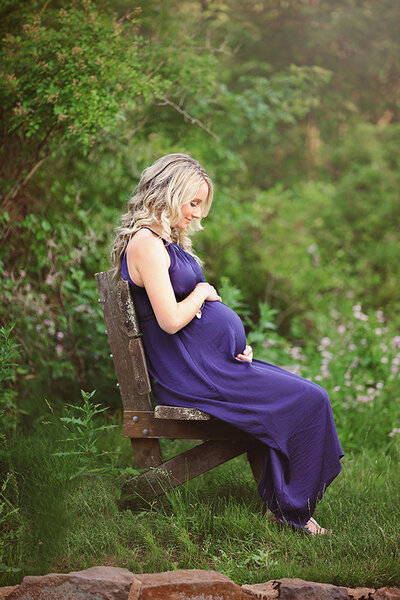 expecting mom wearing a blue dress sitting and holding her belly looking down at her baby to be