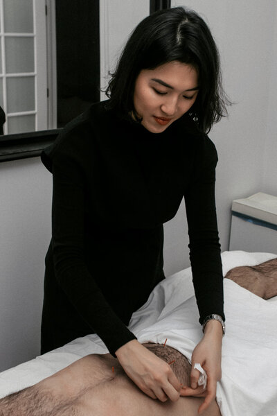 Woman with short black hair dressed in all black administers acupuncture on male patient's lower abdomen.