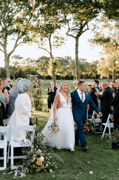 bride and groom exiting outdoor wedding ceremony at sunset while their loved ones cheer at swan lake golf club in manorville - by wedding photographer Daniella Diaz Photo