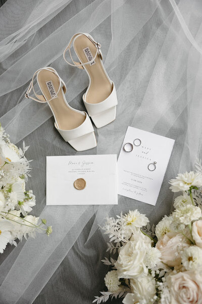 Flat lay featuring a bride’s veil, modern square-toe bridal shoes, and white florals alongside a wedding invitation with a gold wax seal and wedding rings, ideal for wedding stationery inspiration in Ottawa.