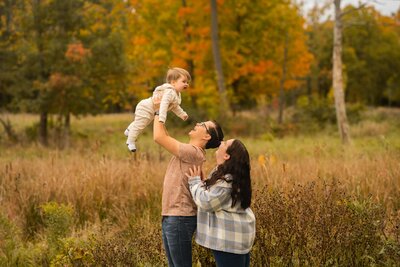mom behind dad who is holding up toddler baby in the fall foliage as they smile up at their baby who is looking around taking in the world 