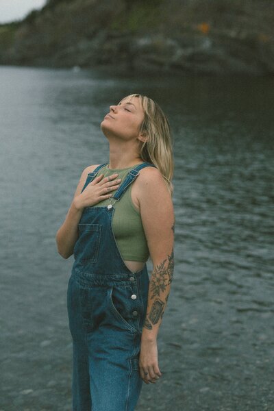 blonde woman with tattoos holds her hand over her heart near the water during Self Love Session