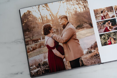 An open photo album featuring a maternity photo of a couple. The woman is wearing a flowing red dress, and the man is in a tan coat. They are standing outdoors at sunset, sharing an intimate moment.