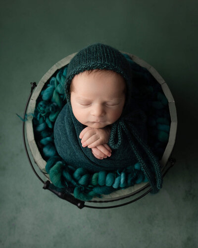 Newborn boy sleeping, wearing dark teal wrap and bonnet, on dark teal background, with hands crossed at chest
