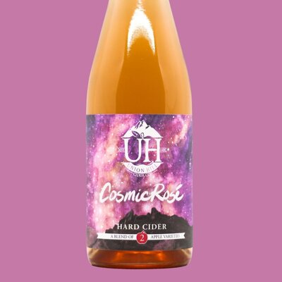 A bottle of Cosmic Rosé hard cider made by Union Hill. Label is bright pink, purple, and yellow like a nebula with Saddle Rock at the bottom of the label.