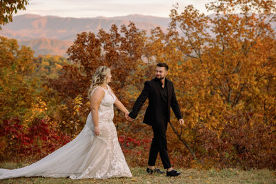 couple elopes to Gatlinburg in the Foothills with the bride and groom holding hands and walking through an outlook in the fall with autumn trees behind them and the Smoky mountains in the distance photographed by photographers in gatlinburg tn