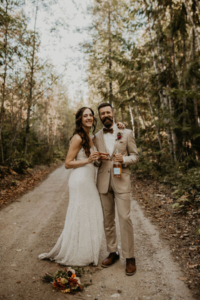 Miranda and her husband, celebrating their wedding in the woods, holding glasses and a bottle, with joyful smiles.