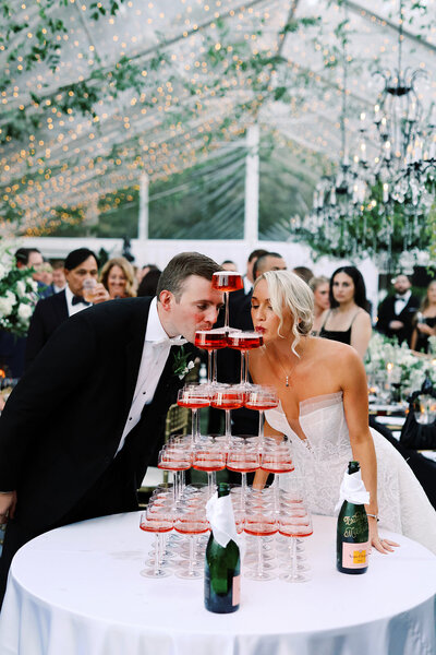Brittany and Michael sip from the champagne tower at their wedding reception at Boxwood Estate. They smile at each other as they take a moment to celebrate, surrounded by elegantly stacked glasses filled with champagne. The tower sparkles under the soft lighting, and their guests cheer in the background, adding to the festive and joyful atmosphere of the occasion.
