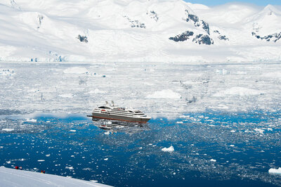 Expedition cruise ship sailing through icy waters surrounded by snow-covered mountains in Antarctica.