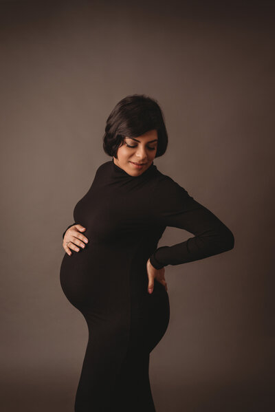 Maternity photos in Marietta GA with Maternity and Newborn Studio of Atlanta by Casey McMinn Photography showcasing a 30 week pregnant mom to be wearing a black pregnancy gown holding her baby bump posing with her hand on her back