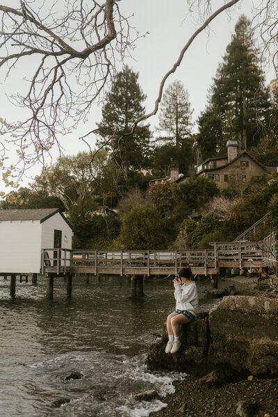 Woman sitting on a coastal rock by the ocean taking a film photo of the water