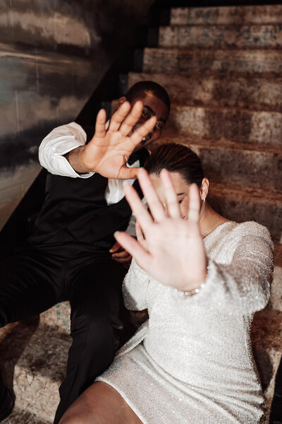 bride and groom hold up their hands to the camera whilst sitting on the stairs at the nomad hotel during their luxury wedding party