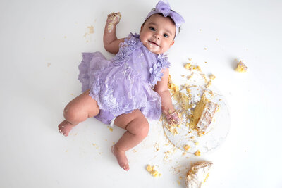 Baby in a lilac lace dress lying on a white floor, holding a fistful of cake with smashed cake scattered around her.