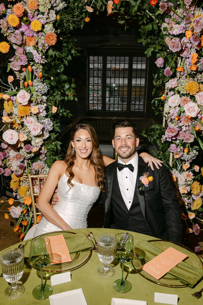 bride and groom sitting in front of colorful lush wedding arch