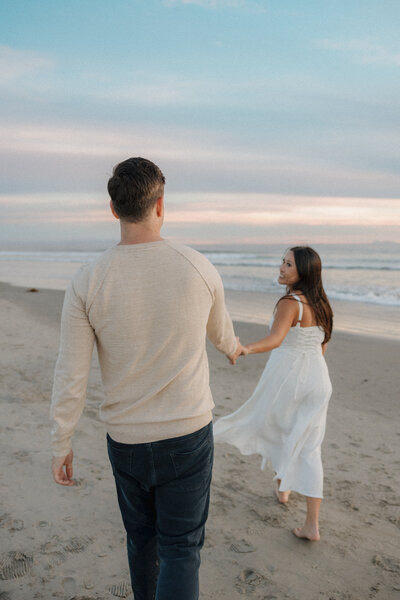 A couple walks hand-in-hand along the shoreline at sunset, the woman looking back with a soft smile.