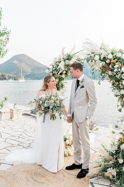 Bride and groom stand hand in hand before a round arch with pastel flowers and pampas on a paved terrace by the sea. In the distance are green hillsides and calm waters with yachts. The bride is wearing a white dress and holding a bouquet and the groom is wearing a light grey suit.
