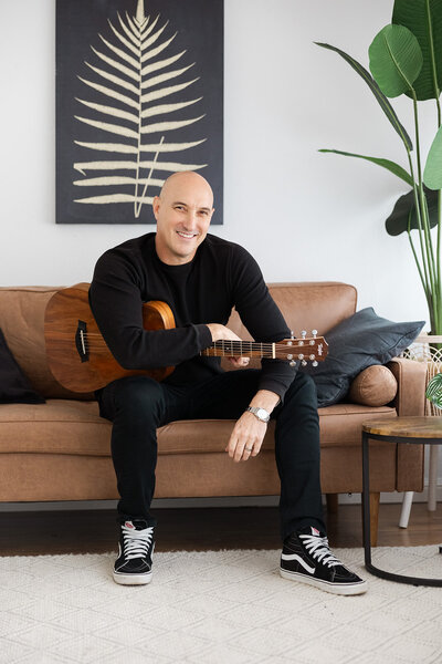 Man sitting on a brown couch wearing all black holding a guitar in a lifestyle Marin brand Photo studio.