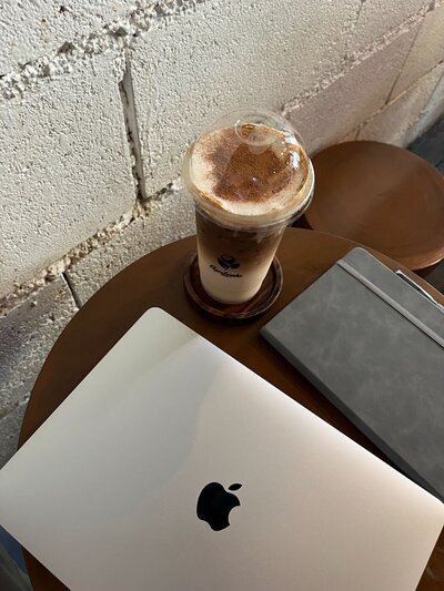 Minimal coffee shop table scene with iced cappuccino in a plastic cup, closed MacBook and grey notebook against a textured white brick wall