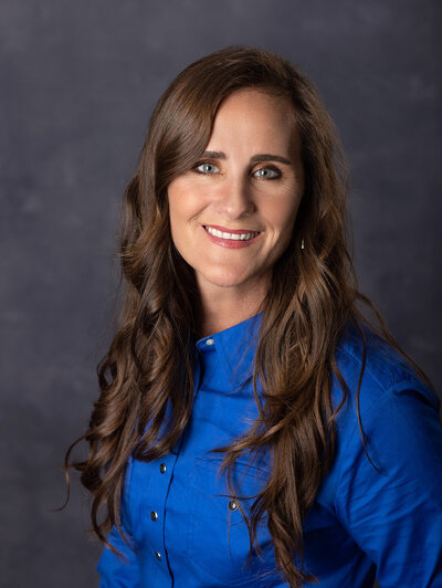 Studio Headshot image of a female professional on a dark gray backdrop wearing a bright vivid blue shirt.