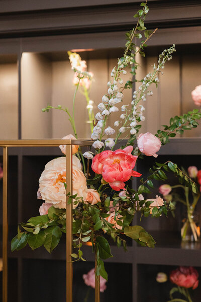 Modern floral installation on a gold stand, featuring garden roses, peonies, and spirea in blush, coral, and ivory hues set against a moody interior backdrop.