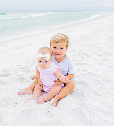 A joyful young boy runs barefoot along the sandy beach, his face beaming with happiness. Dressed in a classic white and yellow romper, he plays near the gentle ocean waves, capturing the carefree essence of childhood in a light and airy family portrait by a Birmingham family photographer."