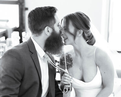 Black and white photo of a Bride and groom at a reception.  Bride has grabbed the groom's tie and pulled him close for a kiss.  He kissed her nose while she is laughing