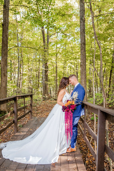 Bride and groom are in the woods on a bridge. He's standing against it in a blue suit and she's facing him with her forehead touching his