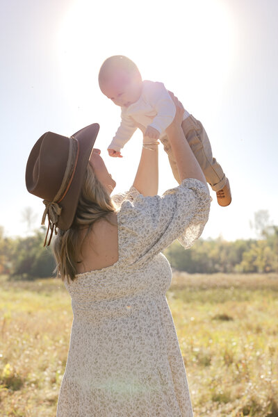 a mother holds her baby up into the sky as the sunrises in a field in Prior Lake, MN