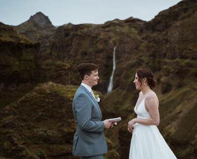 couple is reading their vows in between a waterfall. the bride is smiling as the groom has his vow book out and is reading vows to her. he is in a navy suit and she is in a white dress.