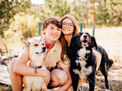 A young couple specializing in Wedding photography in Eau Claire kneels outdoors, smiling at the camera with two dogs—one tan and one black—on a sunny day with trees in the background.